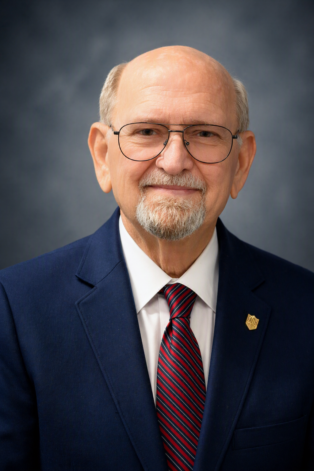 Older man with glasses, white beard, and short hair in a navy suit, white shirt, and red striped tie against a gray background.
