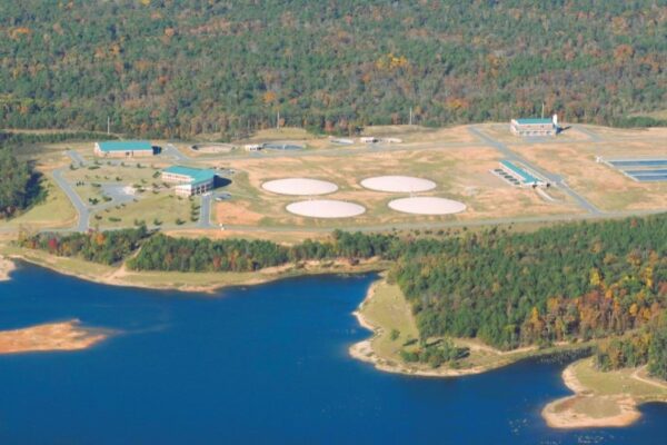 Aerial view of a water treatment facility featuring large circular tanks, multiple buildings, and forested surroundings near water.