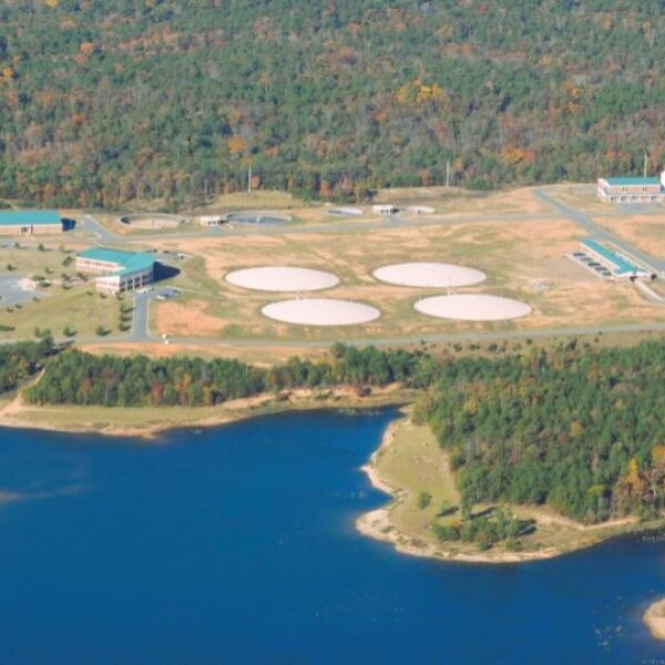 Aerial view of a water treatment facility featuring large circular tanks, multiple buildings, and forested surroundings near water.