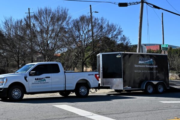A white pickup truck labeled Macon Water Authority Stormwater Management tows a matching black trailer, parked near trees and a sign.