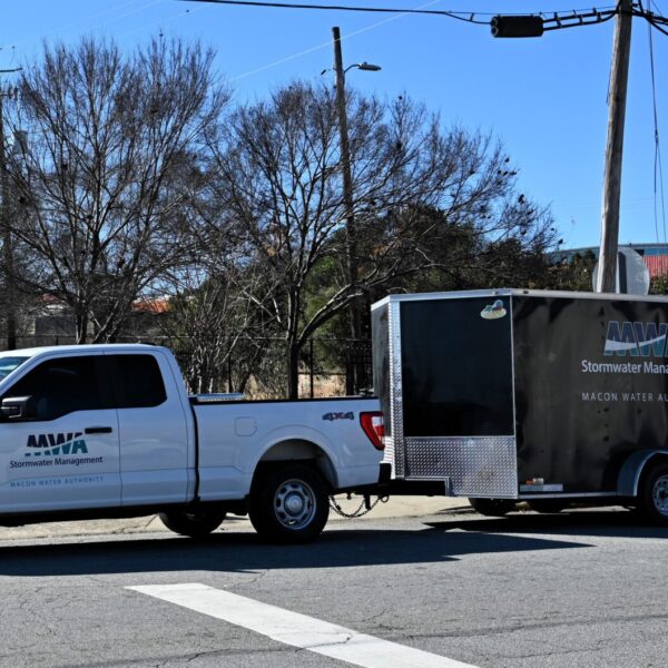 A white pickup truck labeled Macon Water Authority Stormwater Management tows a matching black trailer, parked near trees and a sign.