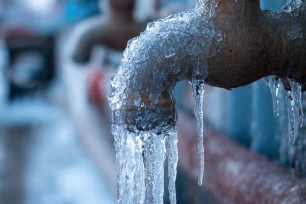 A metal outdoor faucet coated in thick ice and hanging icicles, with blurred frosty pipes visible in the background.