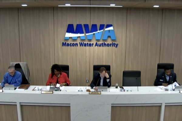 Six people sit at a long conference table under a sign reading Macon Water Authority, reviewing papers in a wood-paneled room.