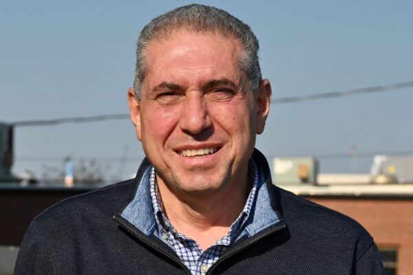 A middle-aged man with short gray hair smiles outdoors in a dark zip-up sweater over a checkered shirt; brick building behind.