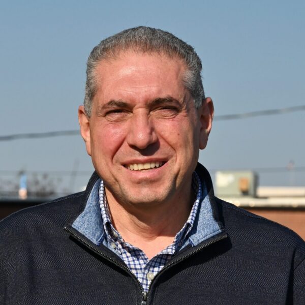 A middle-aged man with short gray hair smiles outdoors in a dark zip-up sweater over a checkered shirt; brick building behind.