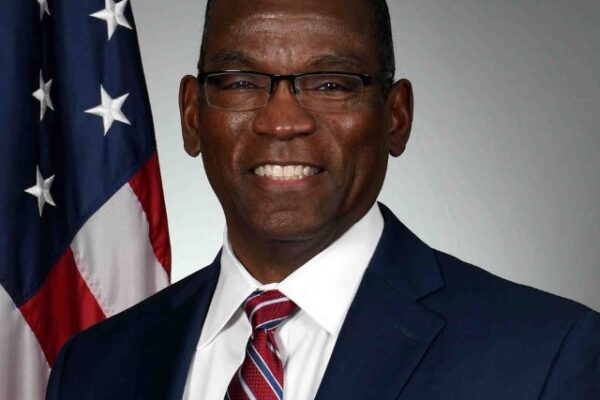 A smiling man in a navy suit, white shirt, and red-striped tie stands before a U.S. flag for a formal portrait on gray backdrop.