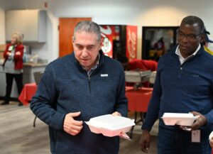 Two men with white takeout containers stand together in a cafeteria, red tablecloths and food trays visible, others in the background.