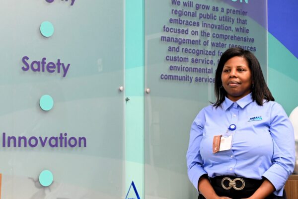 A woman in a blue company shirt stands by a wall displaying Integrity, Safety, Innovation, Stewardship, and a vision statement. A glass award sits nearby.