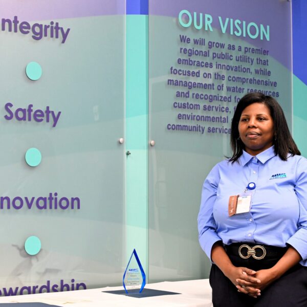 A woman in a blue company shirt stands by a wall displaying Integrity, Safety, Innovation, Stewardship, and a vision statement. A glass award sits nearby.