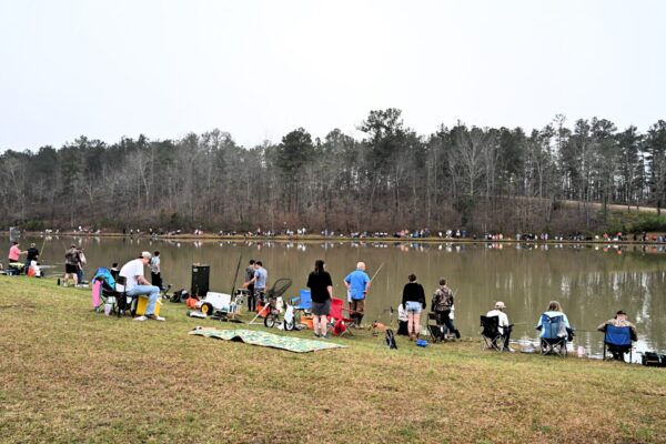 People fish while sitting on chairs and blankets by a pond; more fish opposite. Leafless trees stand under cloudy skies.