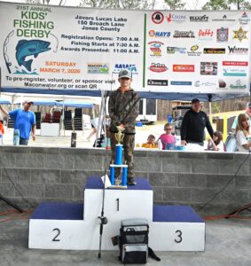 A boy in camouflage stands on a first-place podium with a large blue trophy at a kids’ fishing derby as onlookers watch and a sponsor banner hangs behind.