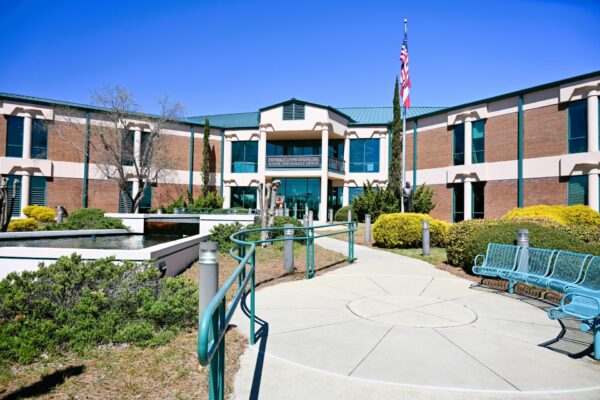 A wide, sunlit walkway approaches a two-story brick building with green trim, American flag, benches, and bushes under blue sky.