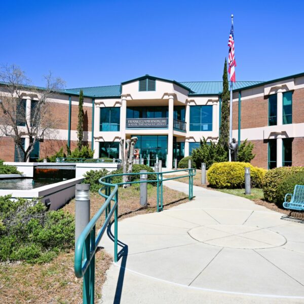 A wide, sunlit walkway approaches a two-story brick building with green trim, American flag, benches, and bushes under blue sky.