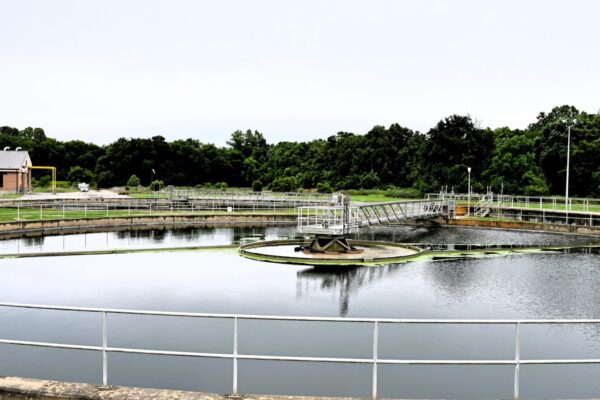 A large circular water treatment basin with metal railings, surrounded by grass, trees, and an industrial building under clouds.