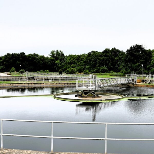 A large circular water treatment basin with metal railings, surrounded by grass, trees, and an industrial building under clouds.
