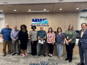 Nine people stand in a row inside an office, smiling at the camera in front of a wall with the Macon Water Authority logo. The group includes men and women in business-casual attire.