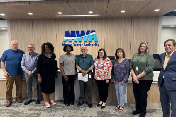 Nine people stand in a row inside an office, smiling at the camera in front of a wall with the Macon Water Authority logo. The group includes men and women in business-casual attire.