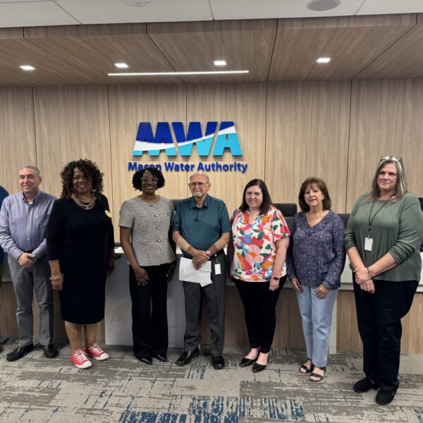 Nine people stand in a row inside an office, smiling at the camera in front of a wall with the Macon Water Authority logo. The group includes men and women in business-casual attire.