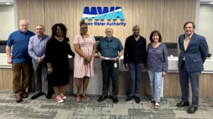 Eight people stand in a row inside an office, smiling at the camera in front of a blue MWA Macon Water Authority sign. A desk and chairs are behind them.