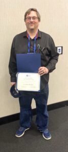 A man stands indoors, smiling and holding an award certificate and blue folder. He wears glasses, black shirt, jeans, and blue shoes.