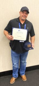 Man in black polo and jeans holding framed certificate and blue hat, wearing gray cap and lanyard, stands by light wall, smiling.