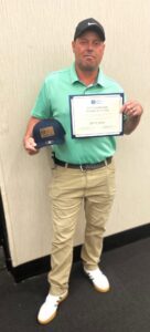 A man in a black cap, green polo, and khaki pants stands indoors on a dark floor, smiling and holding a certificate and baseball cap.
