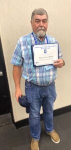 A gray-haired man with a beard, in a blue plaid shirt and jeans, holds a navy cap and Georgia Water Authority certificate indoors, smiling.