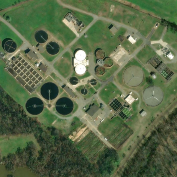 Aerial view of a water treatment facility featuring circular tanks, rectangular basins, and buildings amid grassy fields and woods.