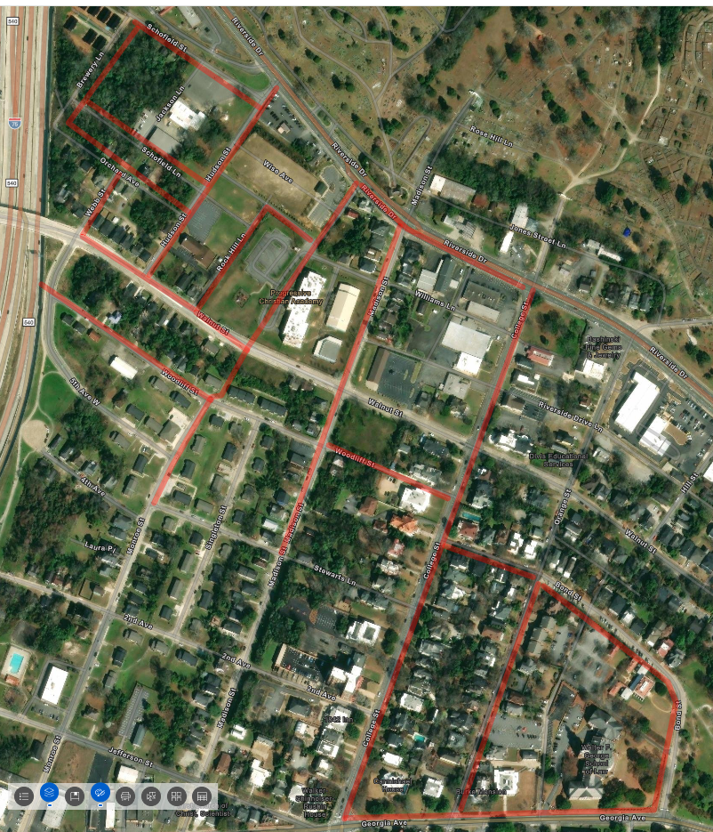 Satellite map of a town with several city blocks outlined in red, showing roads, buildings, and green spaces inside and nearby.