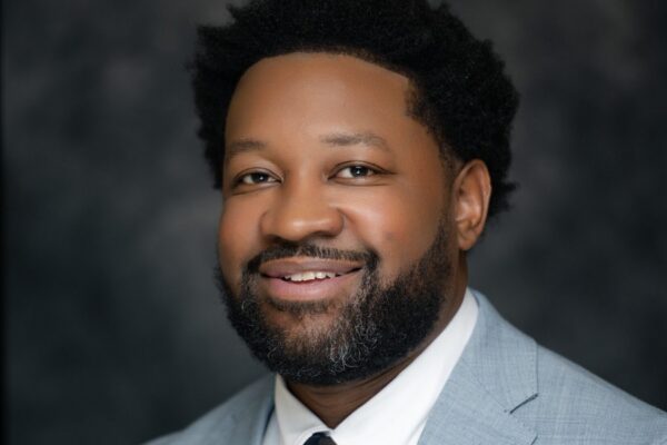 A bearded man with short curly hair smiles in a light gray suit, white shirt, and navy polka dot tie with a tie bar, dark blurred background.