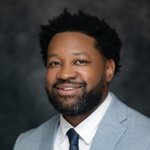 A bearded man with short curly hair smiles in a light gray suit, white shirt, and navy polka dot tie with a tie bar, dark blurred background.