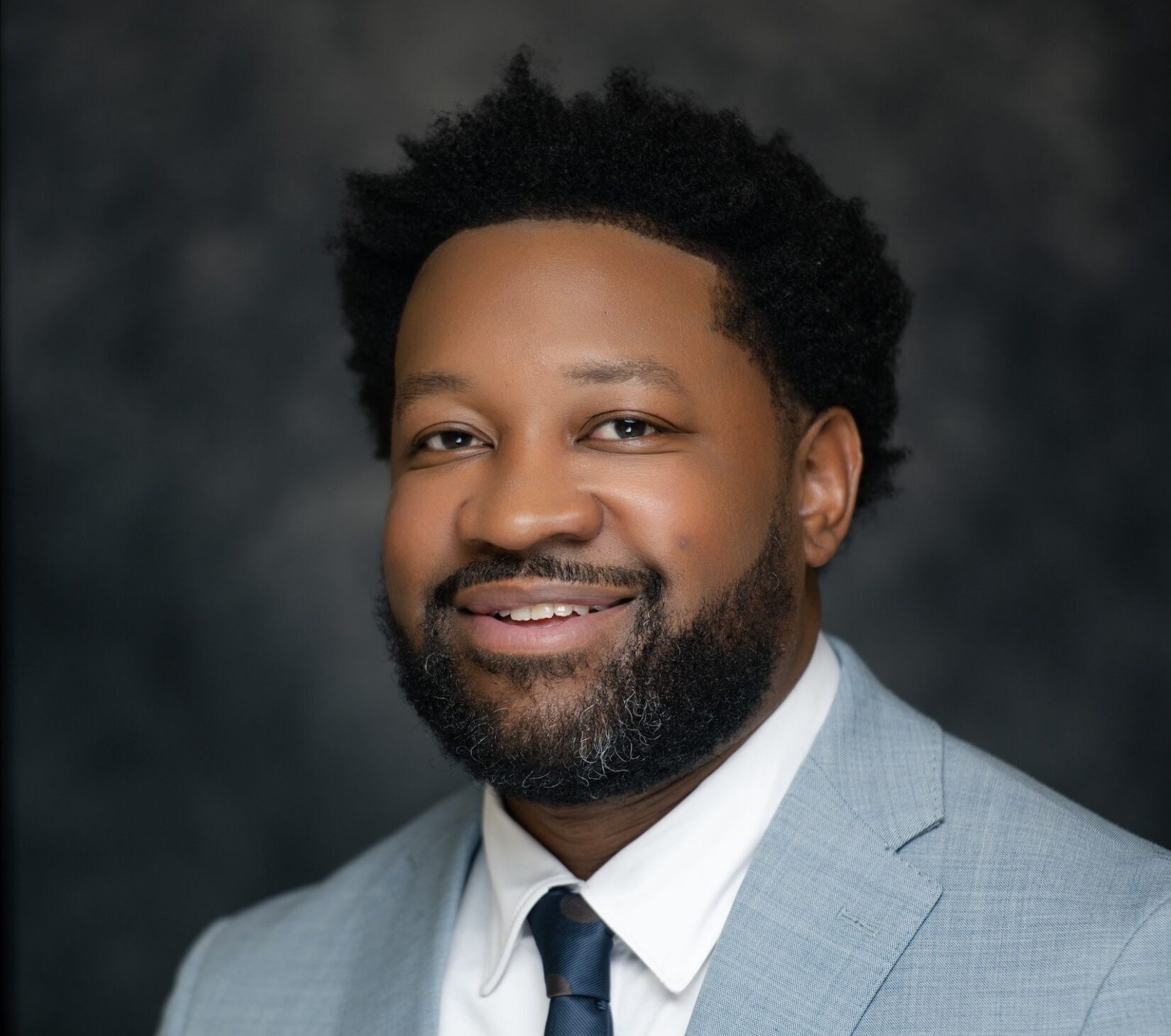 A bearded man with short curly hair smiles in a light gray suit, white shirt, and navy polka dot tie with a tie bar, dark blurred background.