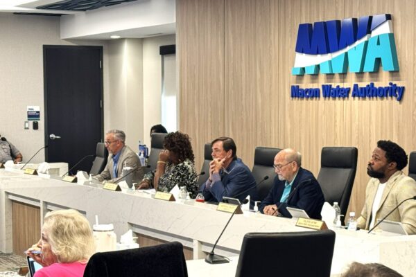 Six people sit at a long conference table under a sign reading Macon Water Authority, with microphones and nameplates. An audience is visible in the foreground.