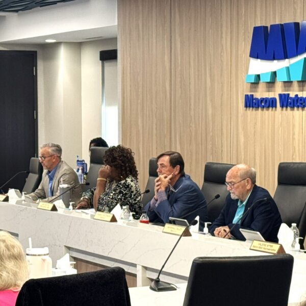 Six people sit at a long conference table under a sign reading Macon Water Authority, with microphones and nameplates. An audience is visible in the foreground.
