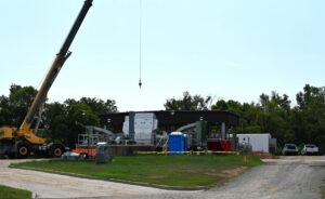 A construction site featuring a crane, building materials, portable toilets, and several vehicles parked nearby. In the background are trees and a clear sky. Part of the site is surrounded by yellow caution tape.