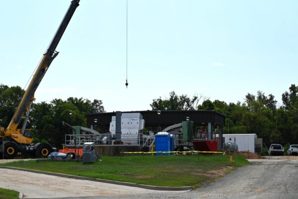 A construction site featuring a crane, building materials, portable toilets, and several vehicles parked nearby. In the background are trees and a clear sky. Part of the site is surrounded by yellow caution tape.