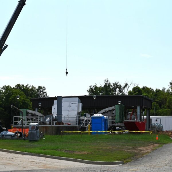 A construction site featuring a crane, building materials, portable toilets, and several vehicles parked nearby. In the background are trees and a clear sky. Part of the site is surrounded by yellow caution tape.
