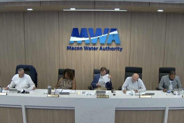 Seven people sit at a long desk under a large Macon Water Authority sign, reviewing documents at a public meeting. The room has wood paneling and microphones on the desk.
