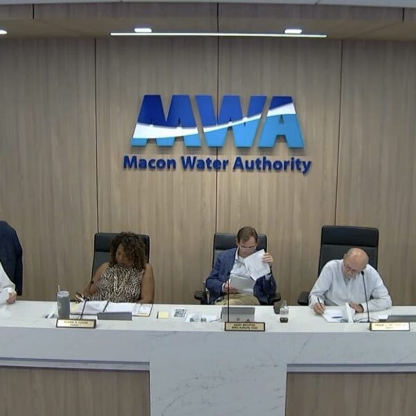 Seven people sit at a long desk under a large Macon Water Authority sign, reviewing documents at a public meeting. The room has wood paneling and microphones on the desk.