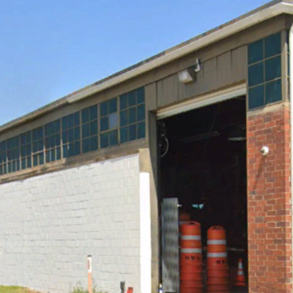 A large industrial building featuring brick and cinder block walls, green-tinted windows, and an open garage door through which orange construction barrels are visible inside.