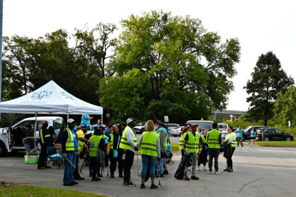 Several people in yellow safety vests stand together beneath a white tent close to parked cars and leafy trees, getting ready for an outdoor cleanup activity.