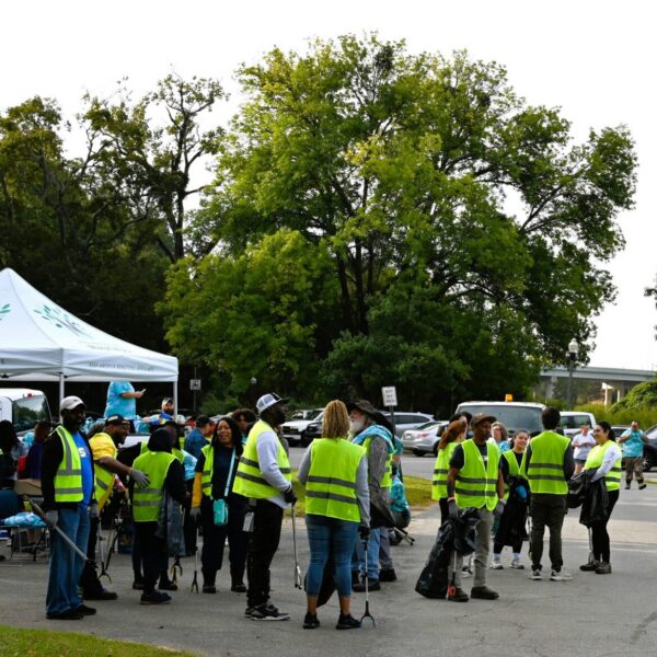 Several people in yellow safety vests stand together beneath a white tent close to parked cars and leafy trees, getting ready for an outdoor cleanup activity.