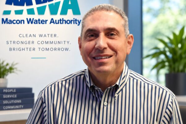 A man in a striped shirt smiles in an office with Macon Water Authority signage. Text reads: Michel Wanna, Executive Director & President. Slogan: Clean water. Stronger community. Brighter tomorrow.
