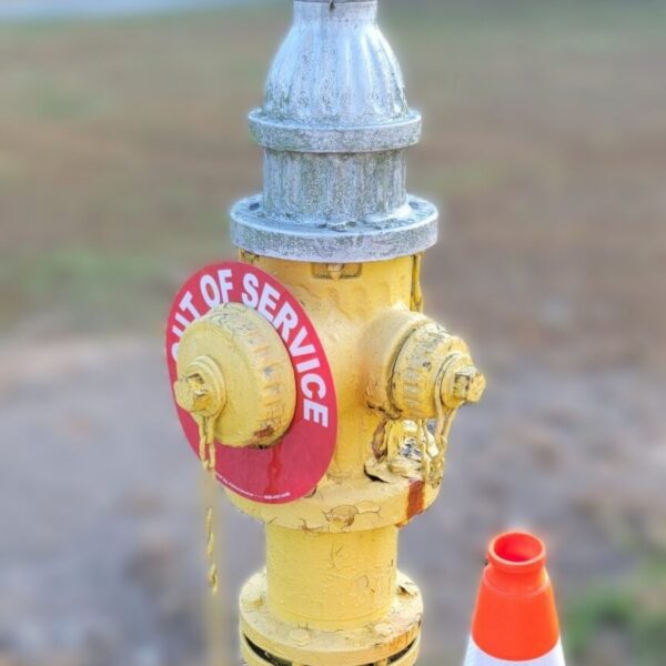 A yellow fire hydrant with a OUT OF SERVICE tag is next to an orange traffic cone. The background is a grassy area and a blurred building.
