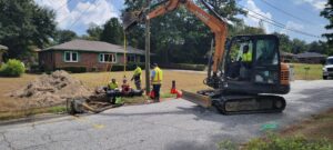 Construction workers operate an excavator to fix underground pipes on a residential street. Dirt piles, tools, and safety cones are scattered nearby, with houses and trees in the background.