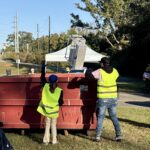 Two volunteers in neon yellow safety vests lift a metal frame into a large red dumpster.