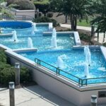 A tiered outdoor fountain with blue pools and several vertical water jets surrounded by landscaping.