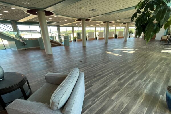 An open, spacious lobby with wood-patterned flooring, several structural columns, and large windows looking out onto a wooded area.