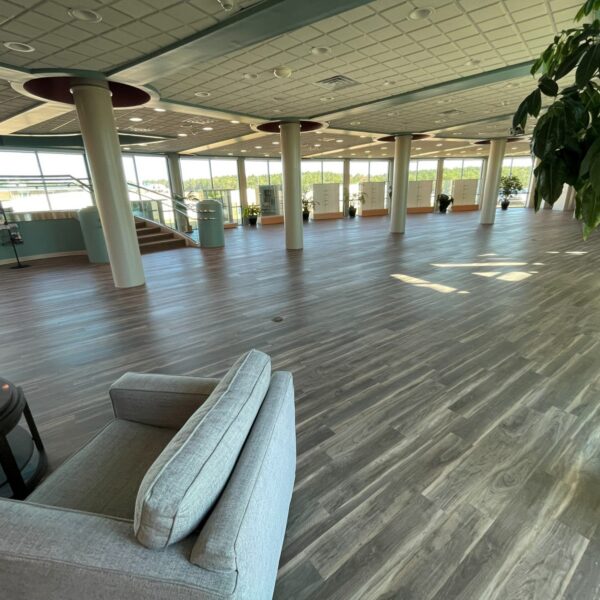 An open, spacious lobby with wood-patterned flooring, several structural columns, and large windows looking out onto a wooded area.