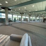 A spacious, carpeted lobby featuring large support pillars, floor-to-ceiling windows, and several people walking through the area.